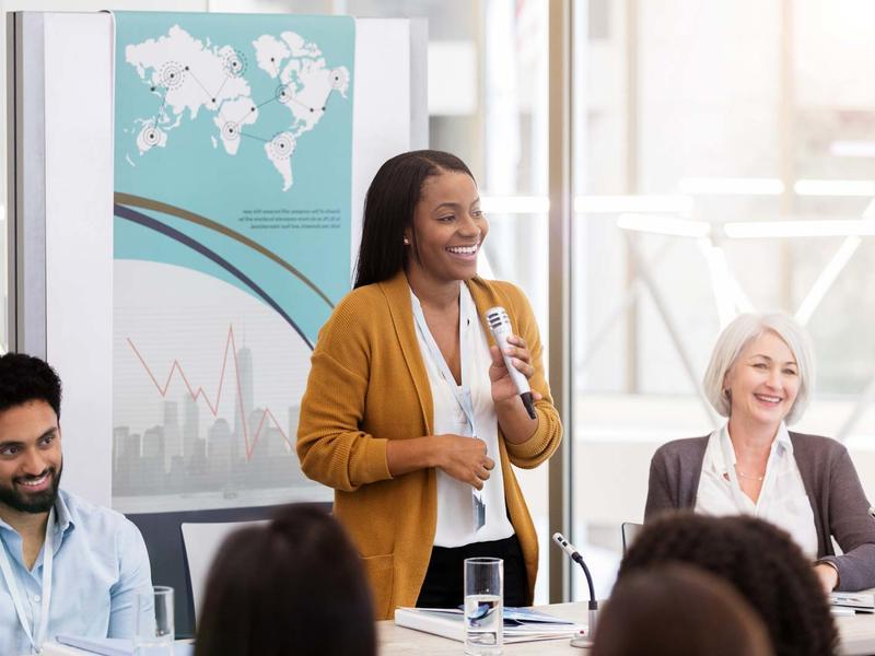 A Black woman with a microphone speaking at a panel, 2 other speakers seated